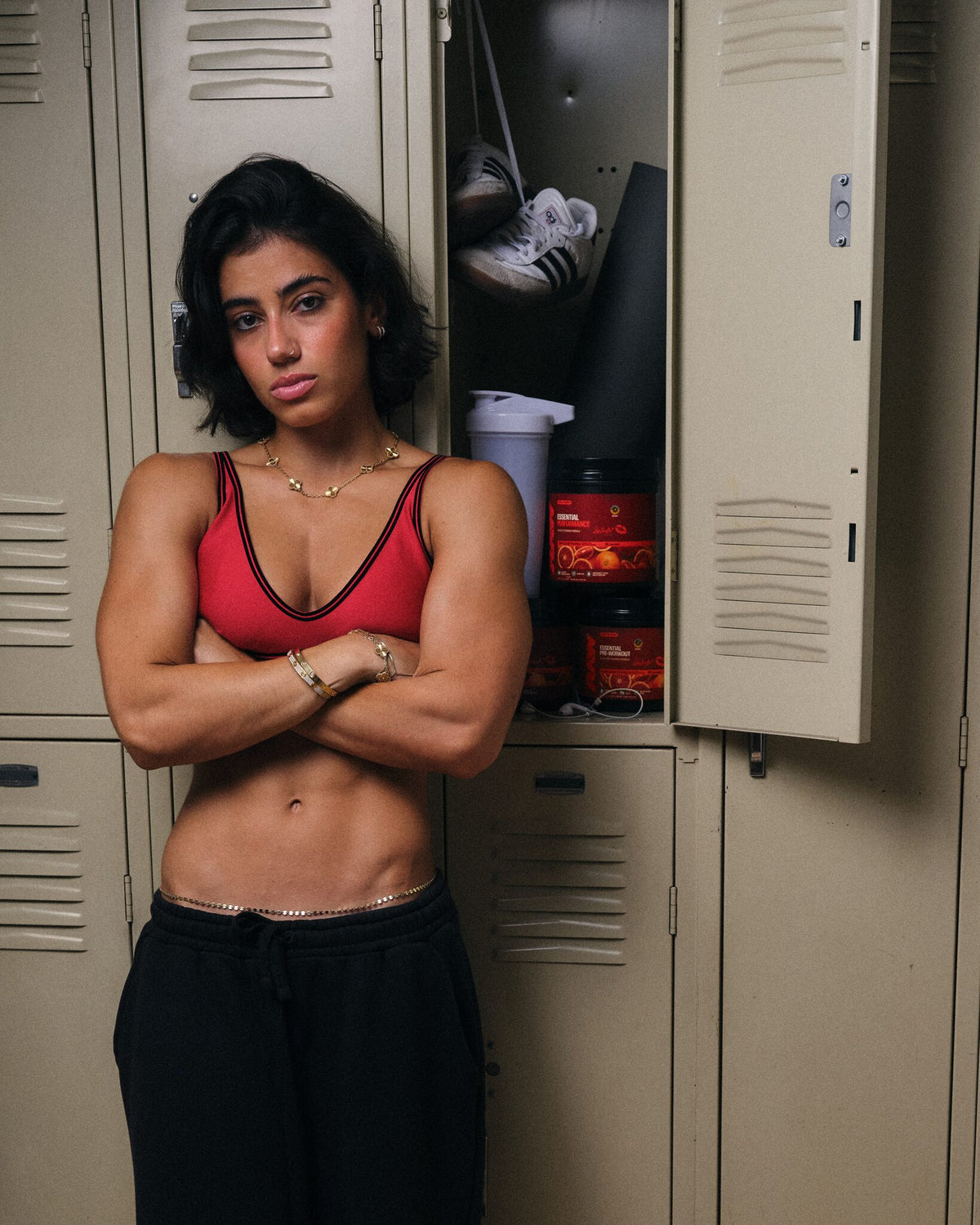 Sara Saffari in front of an open gym locker holding her RAW Sour Blood Orange Essential Performance and Essential Pre-Workout tubs