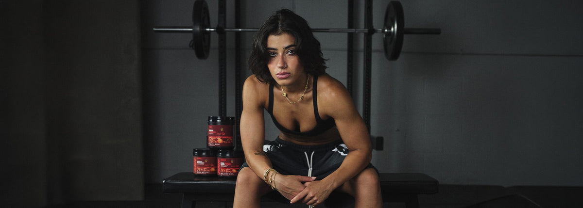 Sara Saffari seated on a gym bench with her RAW Sour Blood Orange Essential Pre-Workout, Essential Performance, and EAA Plus tubs behind her