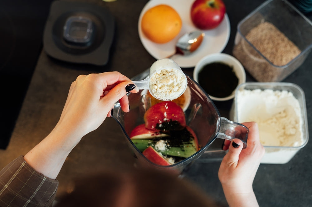 younf womans hand pouring protein powder into blender with fruit and vegetables