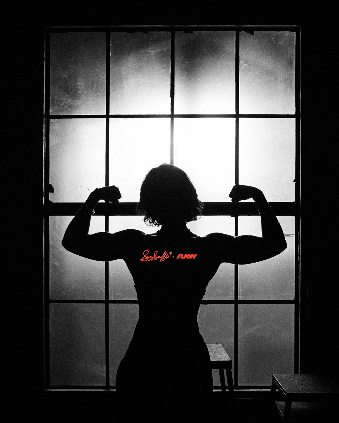 Sara Saffari silhouetted in front of a gym window, flexing, with the Saffari x RAW logo on her back