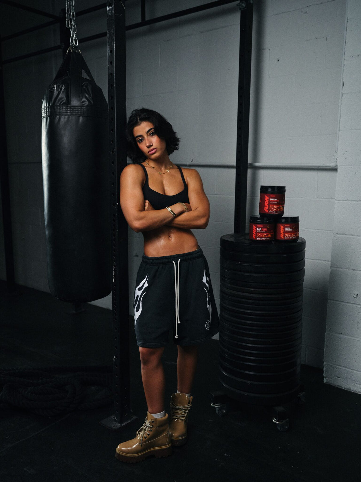 Sara Saffari standing beside a punching bag with her RAW Sour Blood Orange Essential Performance, EAA Plus, and Essential Pre-Workout tubs stacked nearby